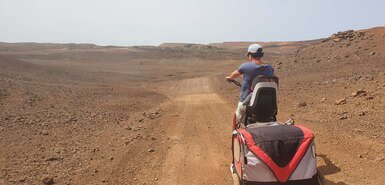 Frau mit Fahrrad, Kindersitz und Anhänger fährt durch eine wüstenartige Landschaft